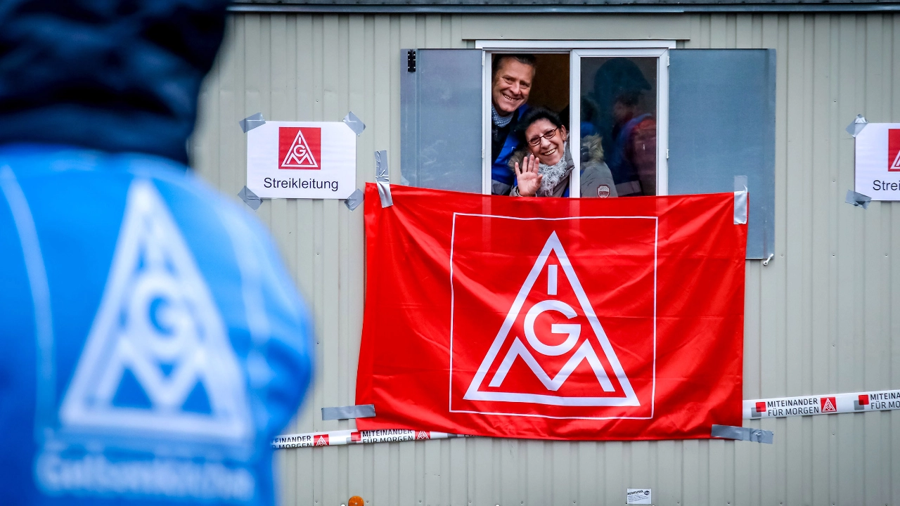 Eine Frau und ein Mann winken aus einem Bauwagen an dem eine IG Metall-Fahne hängt.