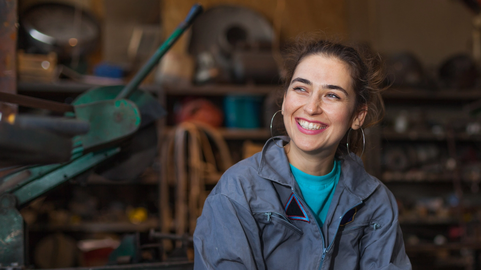 Junge Frau im Blaumann sitzt lächelnd in einer Werkstatt.