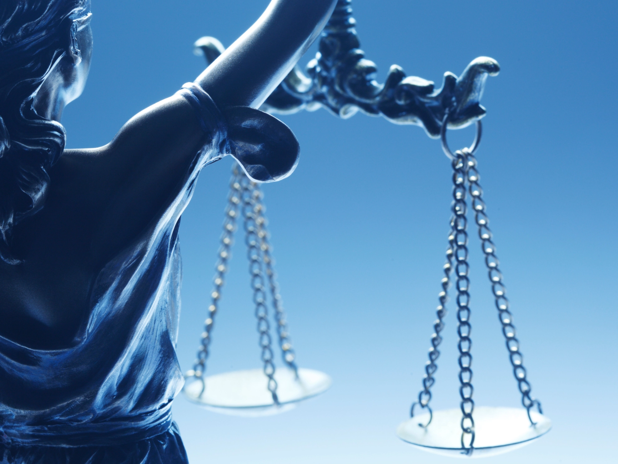 Ein Bild von Justizia als Bronzestatue vor blassblauem Hintergrund, auf dem sie eine Waage hält