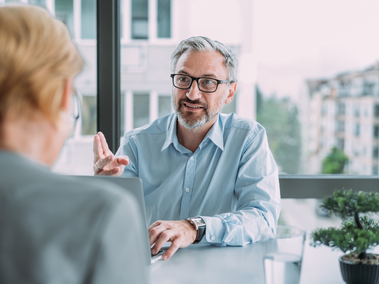 Ein Mann am Schreibtisch hinter einem Laptop im Gespräch mit einer gegenübersitzenden Frau.